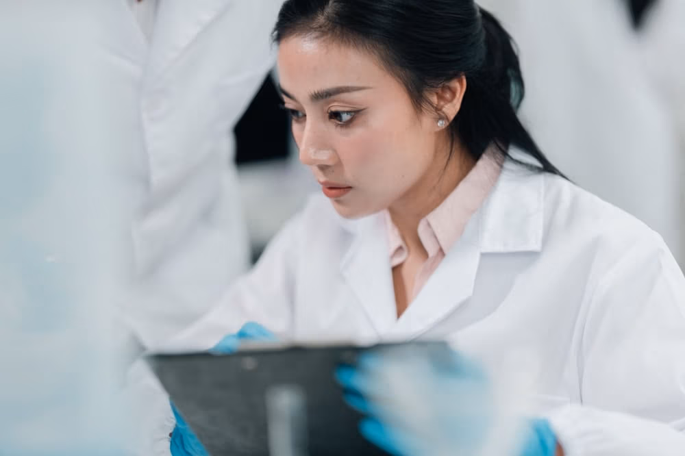 Focused lab technician in white coat and gloves reviewing same day test results on a clipboard in laboratory setting